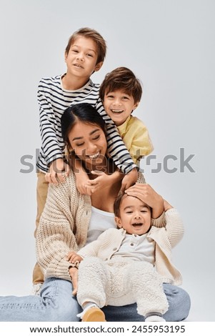 A young Asian mother sits on the floor with her children, sharing a loving embrace in a studio setting against a grey background.