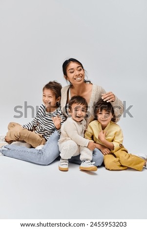 A young Asian mother sits peacefully on the ground with her kids in a studio setting against a grey background.
