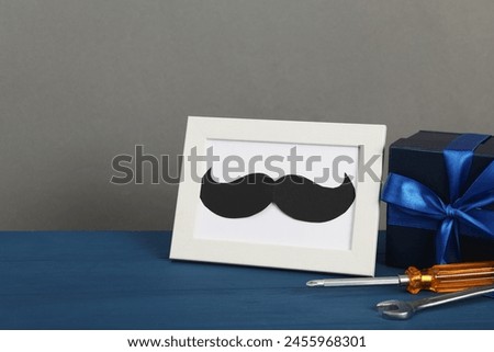 Happy Father's Day. Gift box, tools and frame with mustache on blue wooden table, closeup. Space for text