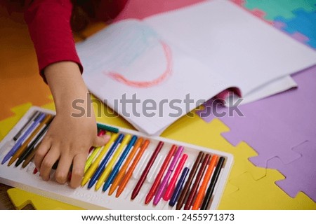 Details on the hands of a little girl taking out colorful pencil from pencil case, drawing beautiful cloud with rainbow, lying on a multi colored puzzle carpet in cozy home interior. Kids education