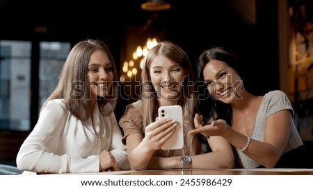 A smiling woman and her beautiful friends in a cafe where they are meeting. A woman shows something on her smartphone. Women discuss each other.