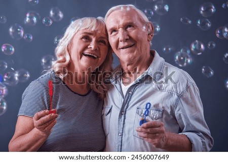 Beautiful old man and woman in casual clothes are blowing soap bubbles, hugging and smiling, on gray background Royalty-Free Stock Photo #2456007649