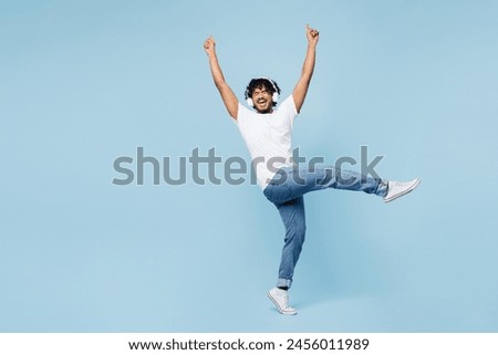 Full body young happy Indian man wear white t-shirt casual clothes listen to music in headphones raise up hands isolated on plain pastel light blue cyan background studio portrait. Lifestyle concept