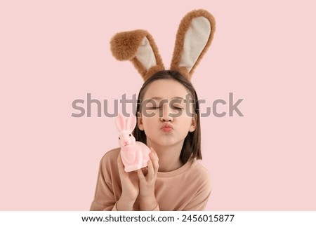 Happy little girl in Easter bunny ears with toy rabbit blowing kiss on pink background