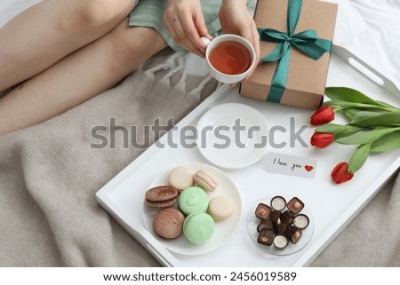 Tasty breakfast served in bed. Woman with tea, desserts, gift box, flowers and I Love You card at home, closeup