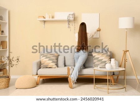 Young woman hanging picture on beige wall in light living room