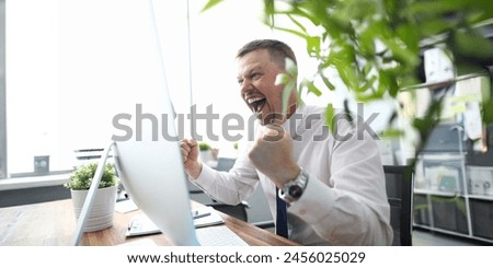 Portrait of cheerful man looking at computer monitor with gladness and making successful gesture. Businessman finishing project and wins big. Blurred background