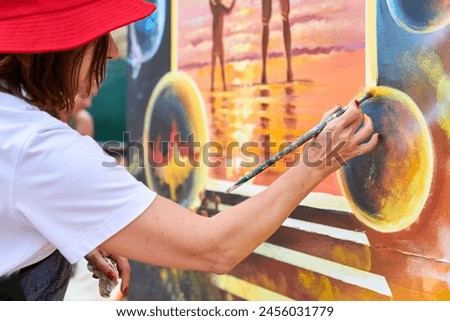 Female painter in red hat draws picture with paintbrush on canvas for outdoor street exhibition, close up side view of female artist apply brushstrokes to canvas, symphony of art creativity
