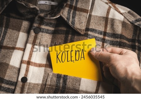 Yellow card with a handwritten inscription "Kolęda", held in the hand against the background of a brown plaid shirt (selective focus), translation: carol