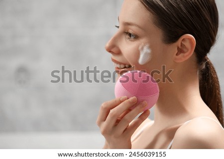 Young woman washing face with brush and cleansing foam indoors. Space for text