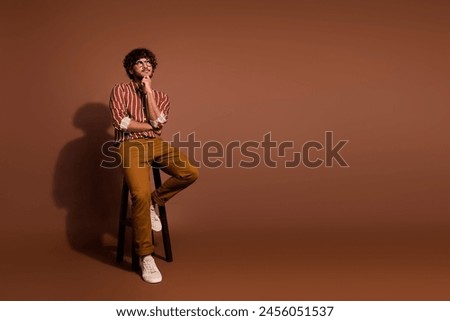 Full size photo of nice young man sit stool look empty space wear striped shirt isolated on brown color background