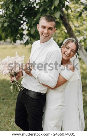 A man and woman are posing for a picture, with the man holding a bouquet of flowers. The woman is wearing a white dress and a veil, and the man is wearing a white shirt and black pants