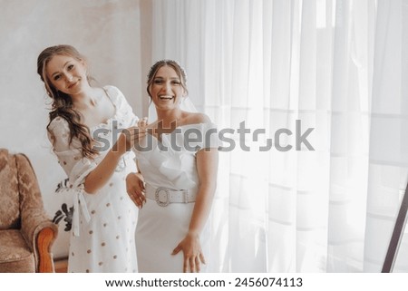 Two women are posing for a picture in a room with a chair and a potted plant. They are both wearing white dresses and smiling