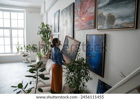 Interested female artist holds painting in hands and searches for suitable place on wall of home to hang result of creative work. Thoughtful designer woman decoration art studio with oil picture. 