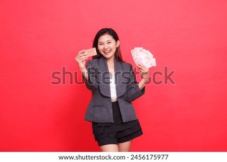 gesture of happy Asian office woman holding credit card and money in front of her, wearing a jacket and skirt on a red background. for financial, business and advertising concepts
