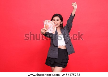 The expression of a cheerful Asian leader woman holds a credit card and money in front of her, clenches her fists and wears a jacket and skirt on a red background. for business and advertising