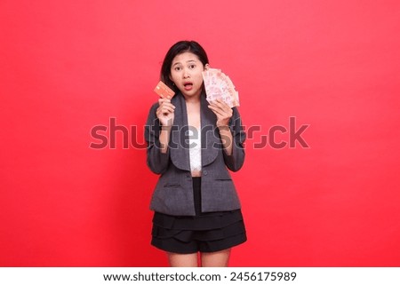 Asian office girl gestures very surprised holding credit card and money in head area wearing jacket and skirt on red background. for transaction, business and advertising concepts