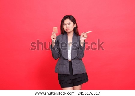 The expression of an indonesia office woman frowning, holding a debit credit card while pointing to the left, wearing a jacket and skirt on a red background. for financial, business and advertising