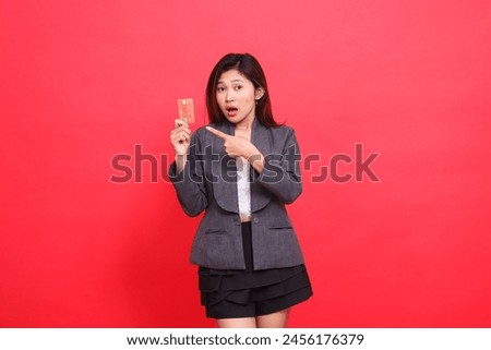 Asian businesswoman's expression shocked at the camera, holding a debit credit card while pointing at it, wearing a jacket and skirt on a red background. for financial, business and advertising