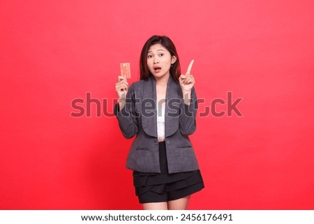 Asian office girl expression shocked at the camera, holding a debit credit card while pointing upwards, wearing a jacket and skirt on a red background. for financial, business and advertising concepts
