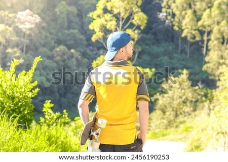 The man takes the longboard by his side, looking towards the horizon. He is seen from behind, wearing a cap, with a forest in the background.
