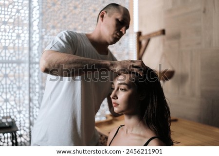 Side view of masseur male doing massage head and hair for young woman client in spa salon. Pretty female getting head massage by focused masseur in wellness center. Concept of rest with spa treatment. Royalty-Free Stock Photo #2456211901