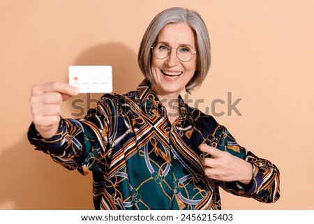 Photo of optimistic pleasant retired person wear print blouse in glasses indicating at debit card isolated on beige color background