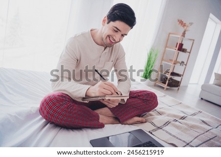Photo of nice cheerful man working from home writing tasks sitting on soft bed in white room indoors
