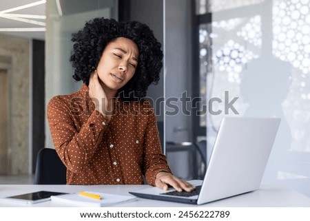 African-American woman experiencing discomfort and neck pain while working on a laptop in a modern office environment, signaling overuse or poor posture. Royalty-Free Stock Photo #2456222789