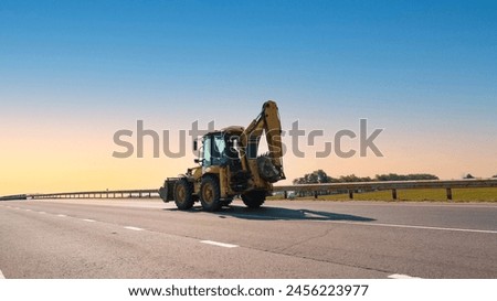 Yellow backhoe loader traveling on highway under clear blue sky signifying ongoing construction work. Open road and sunny day provide perfect setting for heavy machinery operation. Royalty-Free Stock Photo #2456223977