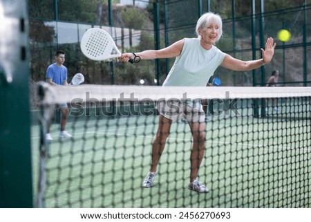 Focused aged woman playing friendly paddleball match on outdoor summer court. Senior people sports concept..