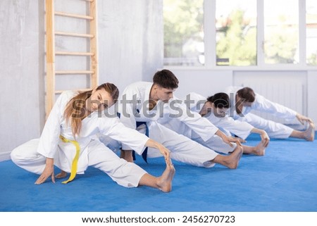 Positive young woman in kimono practicing leg stretching position with others in sports hall