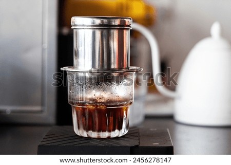 This image shows a Vietnamese drip coffee maker, also known as a Phin, placed on top of a glass filled with dark coffee. The setup is captured on a counter, with a subtle background hinting at a cozy 