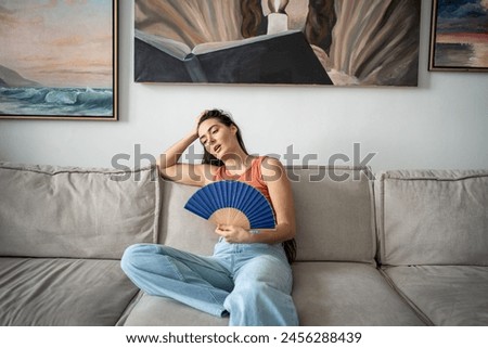 Overheated woman sits on couch in living room at hot summer weather day feel discomfort suffers from heat waving blue fan to cool. Exhausted girl artist sweating cannot working without air conditioner Royalty-Free Stock Photo #2456288439