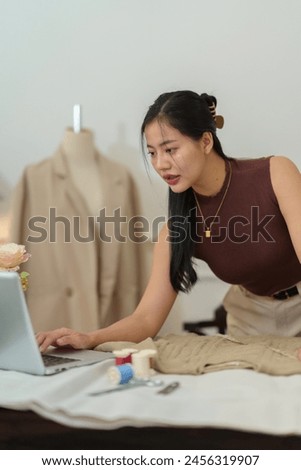 Confident Asian designer woman Beautiful smile using measuring tape for suit jacket with laptop computer in modern studio, clothes hanging on mannequin for customer online sales ordering.