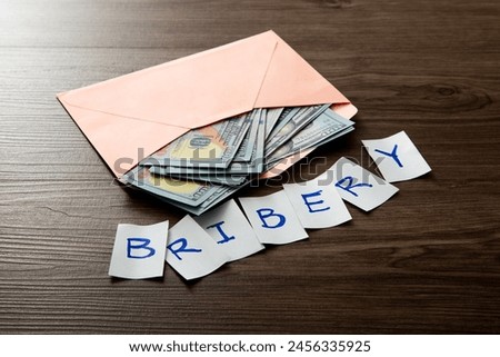 Envelope with us dollar bills and cut-out letters spelling bribery against a dark wood background Royalty-Free Stock Photo #2456335925