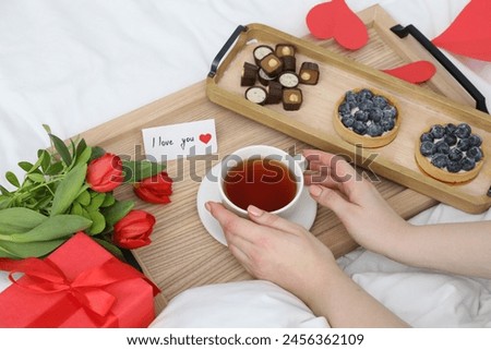 Tasty breakfast served in bed. Woman with tea, desserts, gift box, flowers and I Love You card at home, closeup