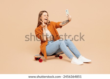 Full body young woman wear orange shirt casual clothes sits on pennyboard skateboard do selfie shot on mobile cell phone show v-sign isolated on plain pastel light beige background. Lifestyle concept