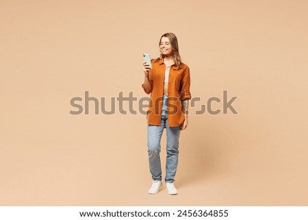 Full body young happy woman she wear orange shirt casual clothes hold in hand use mobile cell phone chatting online isolated on plain pastel light beige background studio portrait. Lifestyle concept