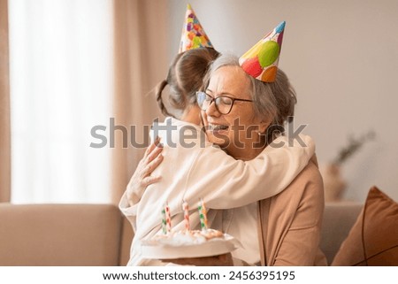 A tender moment captured as an older woman embraces a young girl, both wearing celebratory birthday hats. The affection and joy between them are evident in the heartfelt hug shared. Royalty-Free Stock Photo #2456395195