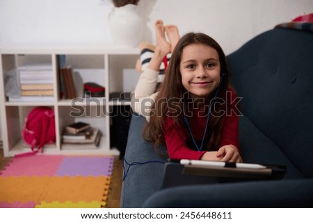 Adorable Caucasian child girl watching cartoons or movie, having fun on digital tablet, lying down on a a comfortable sofa in a cozy home interior. People. Wireless technology. Online communication