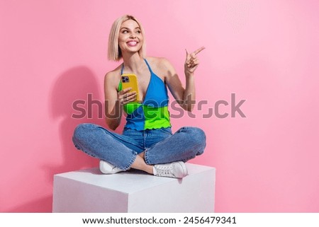 Full size photo of charming woman sit on platform look directing at sale empty space hold smartphone isolated on pink color background