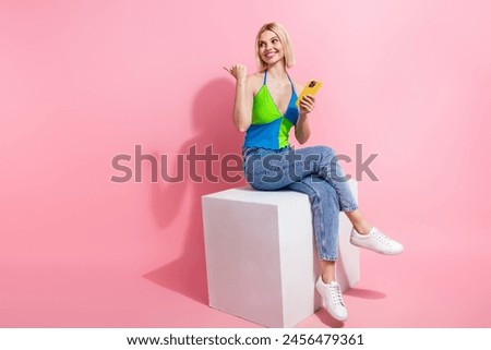 Full size photo of charming woman wear top sit on platform look directing empty space hold smartphone isolated on pink color background
