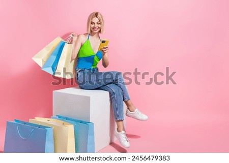 Full size photo of charming woman sit on platform hold clothes look at eshop in smartphone choose outfit isolated on pink color background