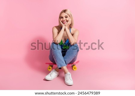 Full size photo of optimistic pretty woman wear colorful top jeans sit on skateboard hands on cheekbones isolated on pink color background