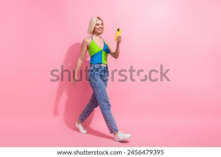 Full size photo of good mood woman wear colorful top jeans look at smartphone walk in empty space isolated on pink color background