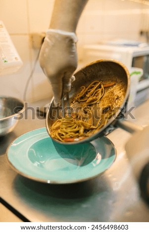 Chef in gloves picks up noodles with metal chopsticks. Professional culinarian skilfully uses special tool serving dish for visitor Royalty-Free Stock Photo #2456498603