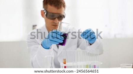 Male chemist holds test tube of glass in his hand overflows a liquid solution of potassium permanganate conducts an analysis reaction takes various versions of reagents using chemical manufacturing. Royalty-Free Stock Photo #2456564651