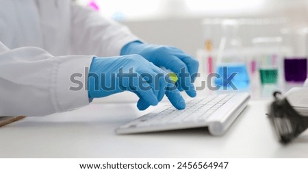 Employee chemical laboratory teacher chemist knocks his fingers keyboard makes notes electronic journal records the analysis data from reactions examining test tubes with the substance arm in gloves. Royalty-Free Stock Photo #2456564947