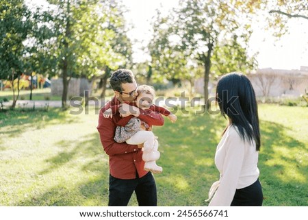 Dad kisses a little girl in his arms next to a standing mom in the park